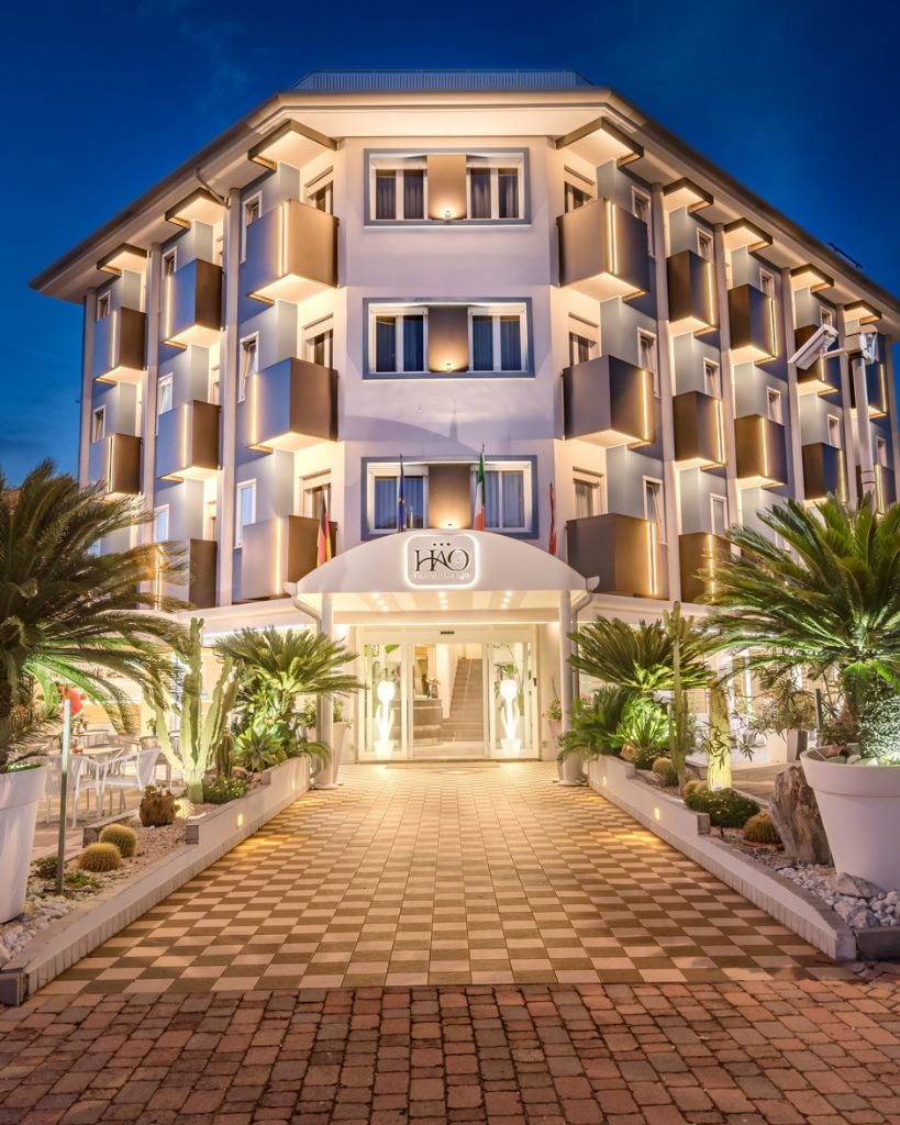Facciata esterna dell'Hotel All'Orologio a Caorle illuminata di notte, con insegna luminosa e design moderno dell'edificio.Facciata esterna dell'Hotel All'Orologio a Caorle illuminata di notte, con insegna luminosa e design moderno dell'edificio.