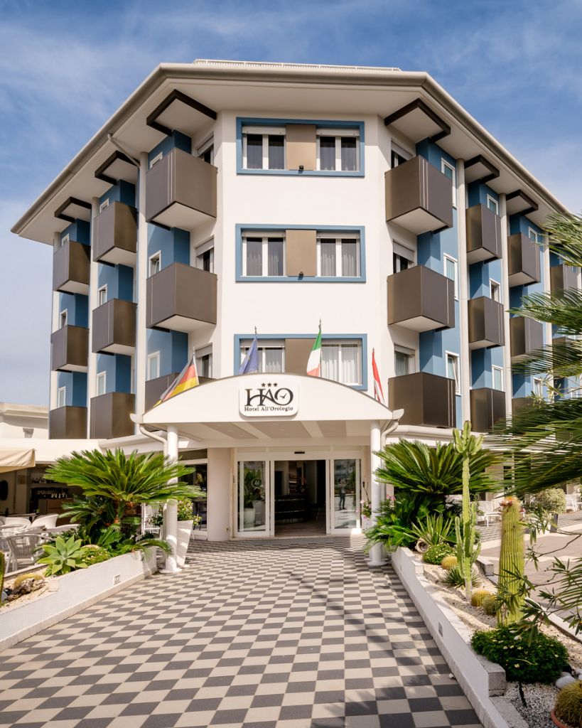 Facciata esterna dell'Hotel All'Orologio a Caorle di giorno, con architettura moderna, balconi e insegna visibile.