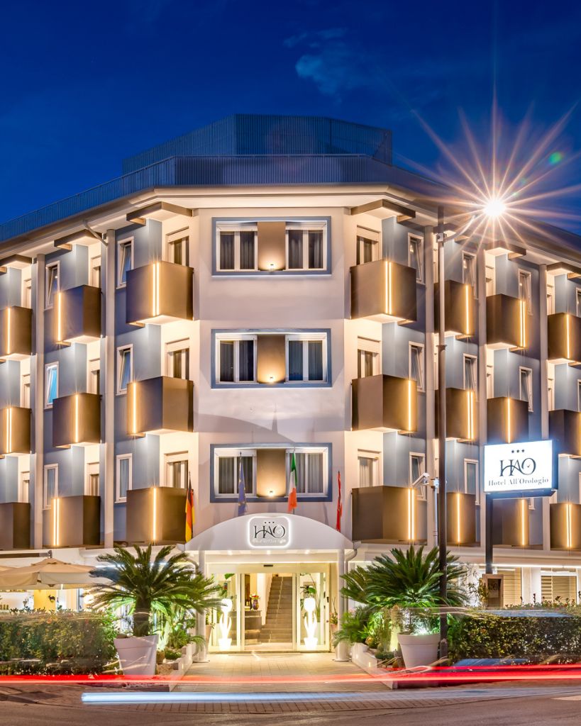 Vista notturna dell'Hotel All'Orologio a Caorle con illuminazione architettonica esterna e insegna luminosa.