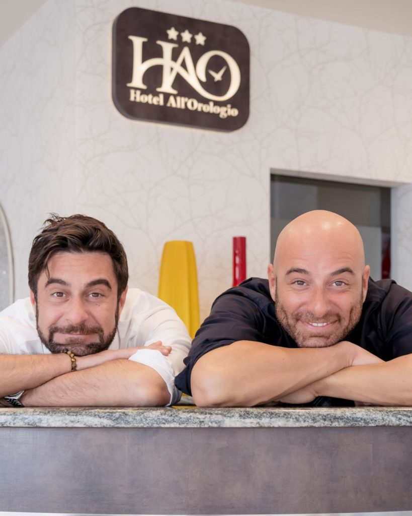 Hotel Caorle All'Orologio Gilberto e Alessio alla reception dell’Hotel All’Orologio di Caorle sorridono appoggiati al banco dell’accoglienza.