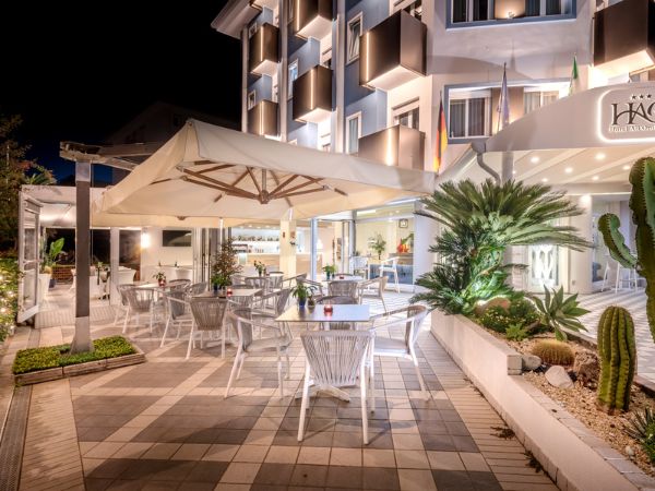 Hotel Caorle All'Orologio Terrazza esterna serale dell'Hotel All'Orologio a Caorle con tavoli, sedie moderne, grandi ombrelloni e piante esotiche illuminati.