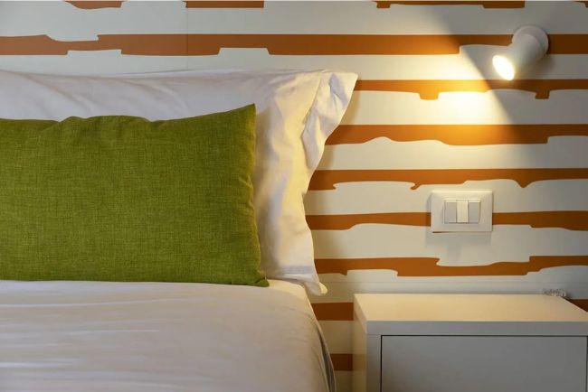 Hotel Caorle All'Orologio Dettaglio letto Camera Papaya con cuscino verde mela, federa bianca e carta da parati a righe arancioni e bianche con lampada da lettura accesa.